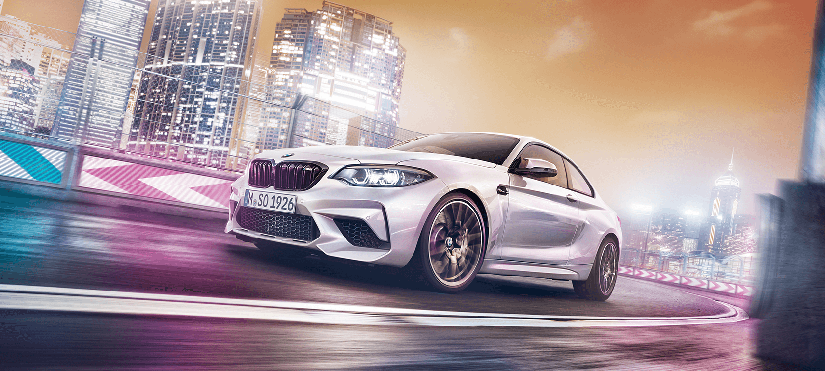 BMW M2 Competition BMW M2 Competition drifting on race track in front of nocturnal cityscape.